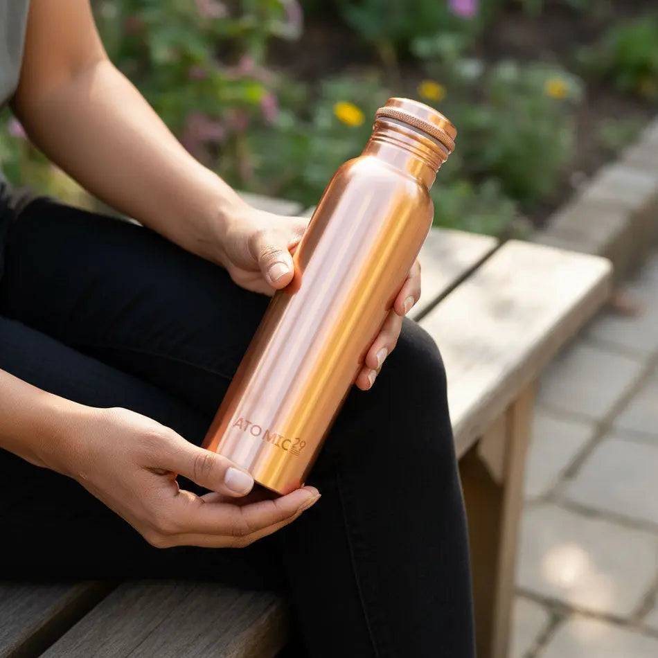 Person holding a copper water bottle with 'Atomic29' branding outdoors.
