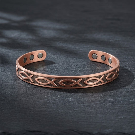 A copper magnetic cuff bracelet with a fish motif, displayed on a dark slate surface.