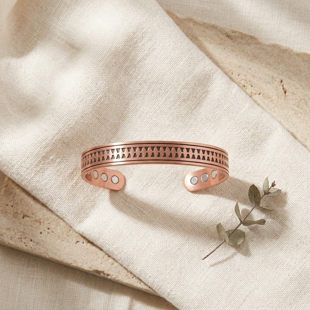 A copper cuff bracelet with a geometric triangle pattern and circular inlay details, displayed on a textured beige linen cloth with a small eucalyptus sprig.