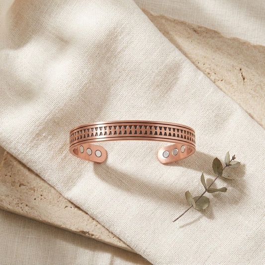 A copper cuff bracelet with a geometric triangle pattern and circular inlay details, displayed on a textured beige linen cloth with a small eucalyptus sprig.