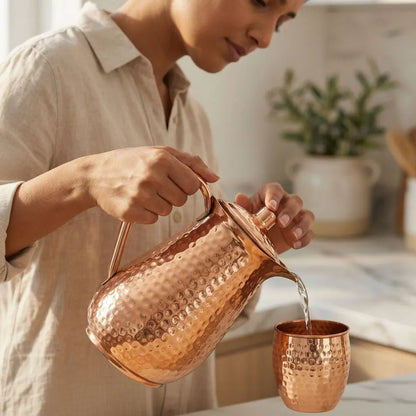 Person pouring water from a copper jug into a matching copper glass in a home setting.
