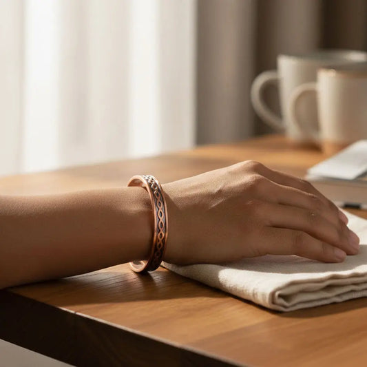 Hand resting on a wooden surface with a copper bracelet, next to a folded napkin and a cup.