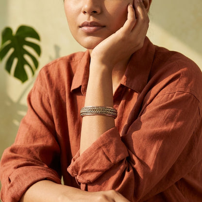 A close-up lifestyle shot of a woman wearing a braided copper bracelet, wearing a rust-coloured linen shirt against a warm beige background with a green leaf shadow.