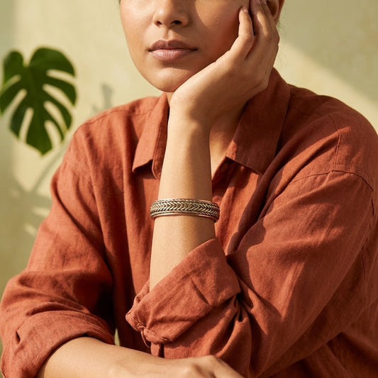 A close-up lifestyle shot of a woman wearing a braided copper bracelet, wearing a rust-coloured linen shirt against a warm beige background with a green leaf shadow.