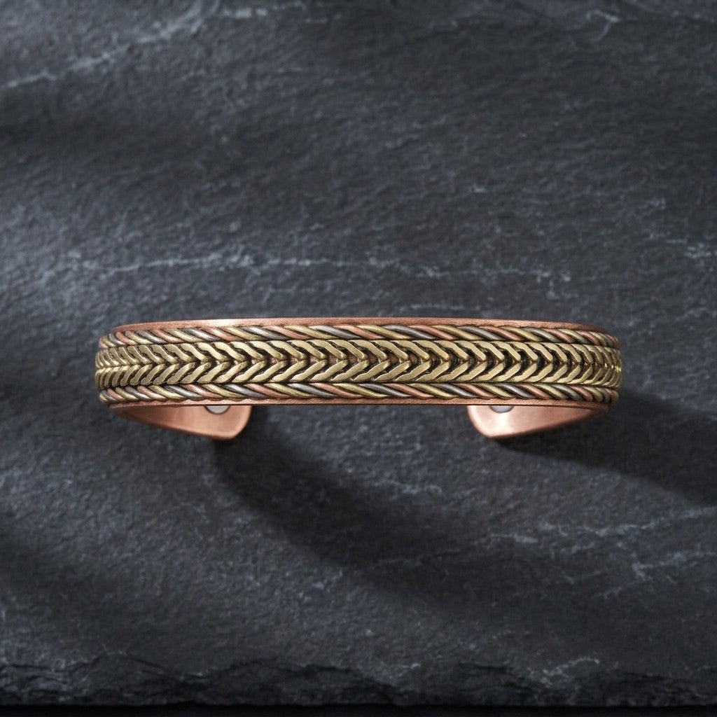 A textured metal cuff bracelet with a woven chevron pattern, featuring a brass toned centre band and copper braided sides, displayed on a dark slate background.