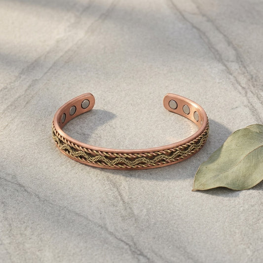 Braided copper magnetic cuff bracelet with a braided copper and brass pattern on the front, displayed on a light grey stone surface next to a dried leaf with three circular magnets on each ends