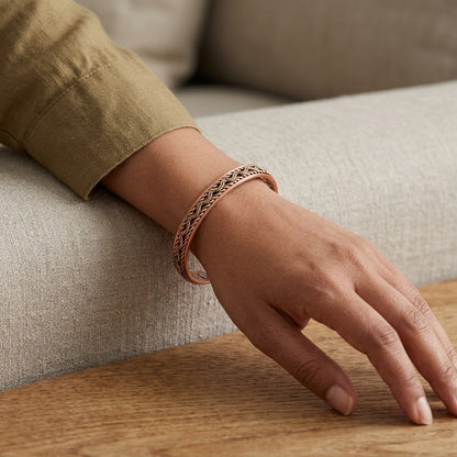 A close-up of a person's hand resting on a wooden table, wearing a braided copper cuff bracelet in a warm rose-gold tone. The person is wearing a khaki-coloured long-sleeve shirt, and the background shows a neutral-toned fabric sofa.