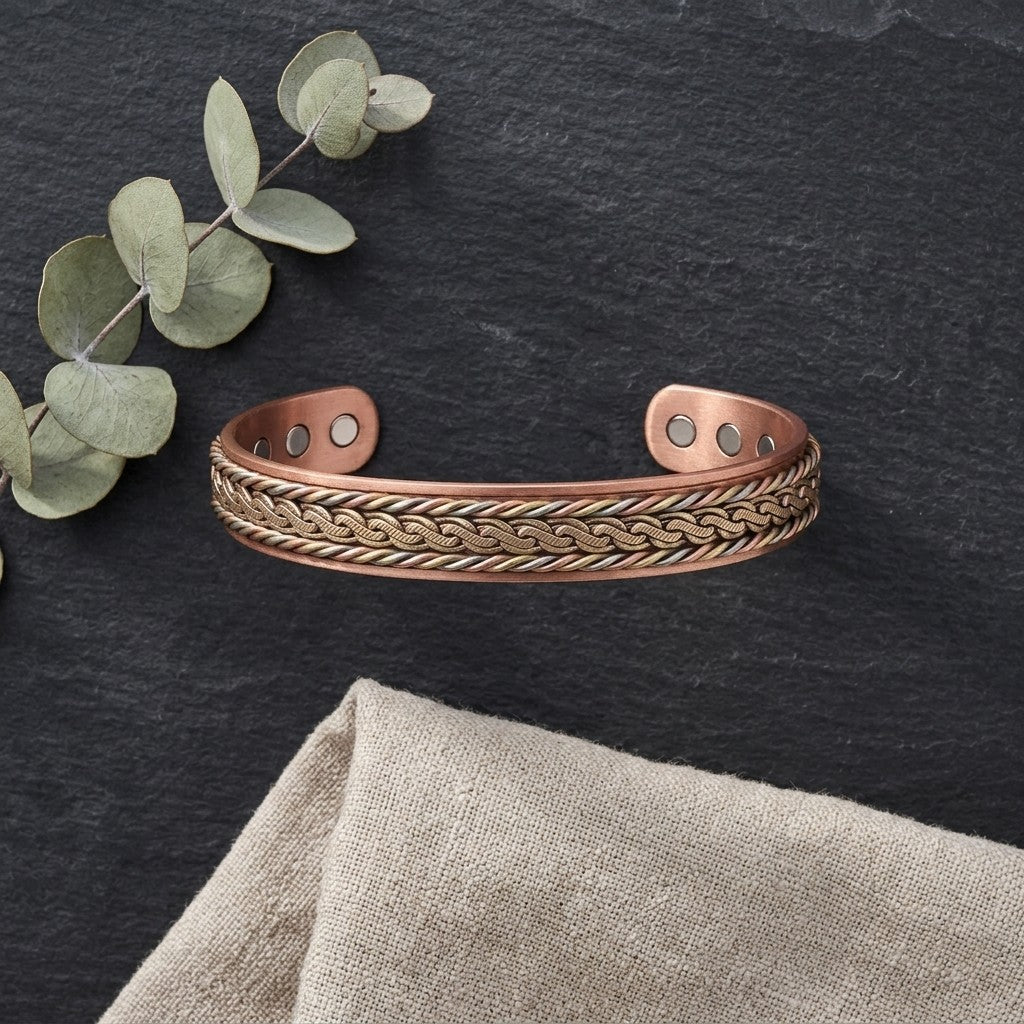 Braided copper magnetic cuff bracelet with a interlock patterned of brass, copper and cupronickel, displayed on a dark slate surface with eucalyptus leaves and a beige fabric napkin.