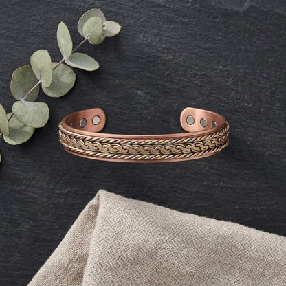 Braided copper magnetic cuff bracelet with a interlock patterned of brass, copper and cupronickel, displayed on a dark slate surface with eucalyptus leaves and a beige fabric napkin.