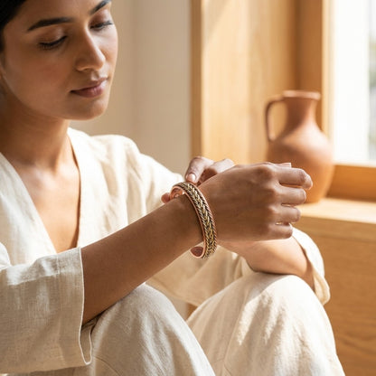 A woman in a cream linen robe sits by a warm window, adjusting a braided copper bracelet on her wrist. A terracotta jug is visible in the softly lit background.