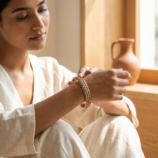A woman in a cream linen robe sits by a warm window, adjusting a braided copper bracelet on her wrist. A terracotta jug is visible in the softly lit background.