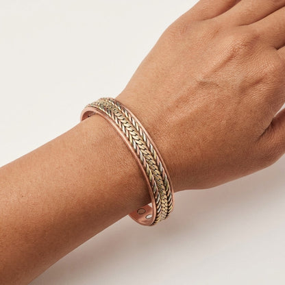 A close-up of a person's wrist wearing a wide cuff bracelet with braided detailing in copper, bronze and cupronickel, set against a clean white background.