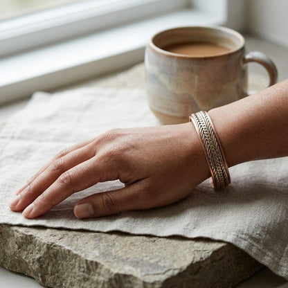 A close-up of a person's hand resting on a textured stone surface next to a ceramic mug, wearing a braided copper magnetic cuff bracelet with woven details.