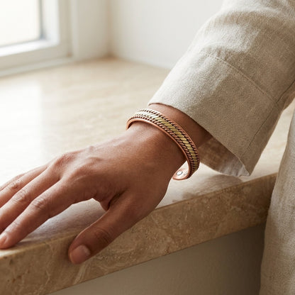 A close-up of a person's hand resting on a stone ledge, wearing a copper cuff bracelet in a warm gold and rose gold tone.