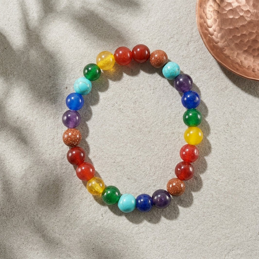 A beaded chakra bracelet with rainbow-colored polished stone beads, arranged in a circle on a textured light grey surface with soft leaf shadows and a copper decorative object in the top right corner.