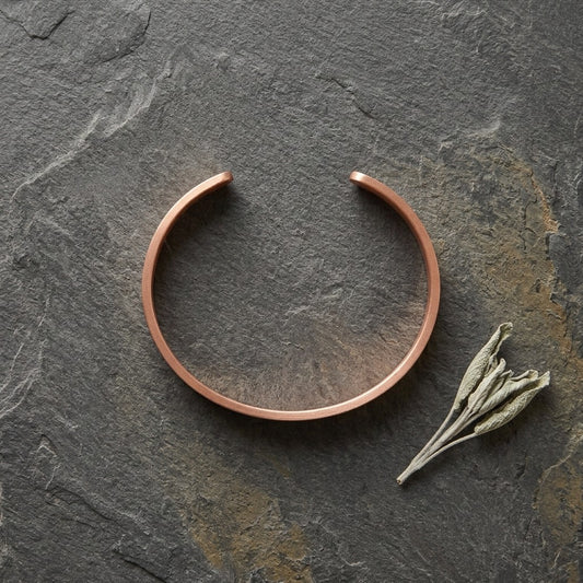 A minimalist copper cuff bracelet is displayed on a dark textured slate surface next to a small dried sprig of sage.