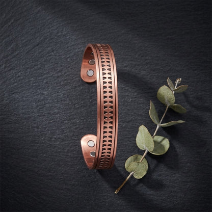 A copper magnetic cuff bracelet with embossed arrow pattern, displayed on a dark slate background next to a small green eucalyptus sprig.