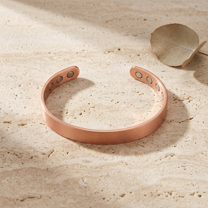 A smooth polish copper bracelet with four circular magnetic accents on each inner ends, displayed on a textured beige stone surface next to a dried leaf.