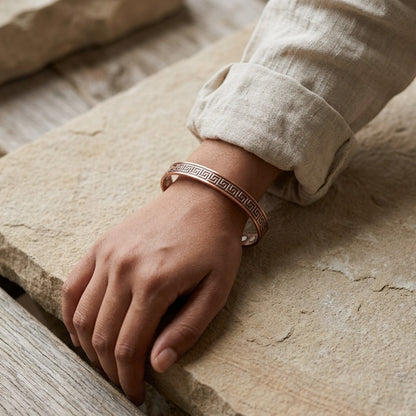 A close-up of a person's hand resting on a rustic stone surface, wearing a copper cuff bracelet with a Greek key pattern.