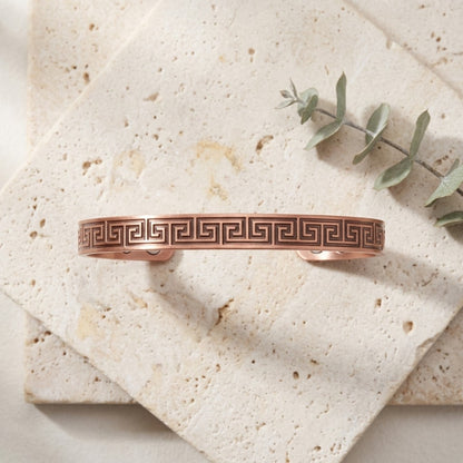 A copper cuff bracelet with a Greek key pattern, displayed on stacked beige stone tiles with a sprig of eucalyptus.