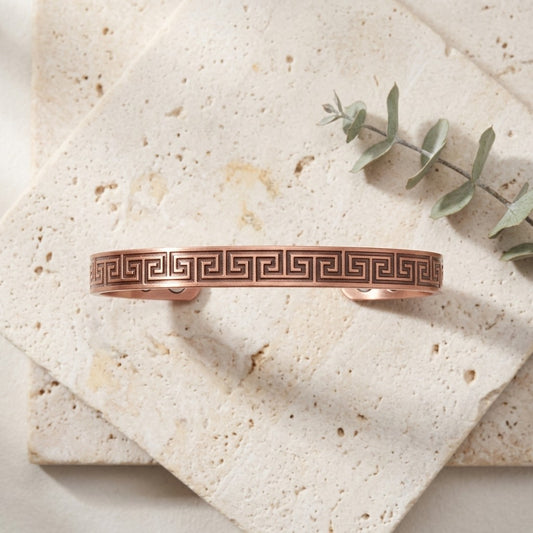 A copper cuff bracelet with a Greek key pattern, displayed on stacked beige stone tiles with a sprig of eucalyptus.