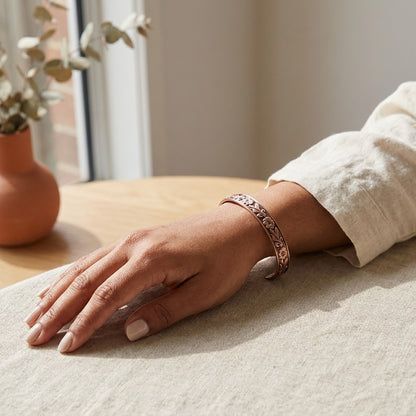 A person's hand resting on a light beige fabric surface, wearing a copper bracelet with an intricate floral pattern.