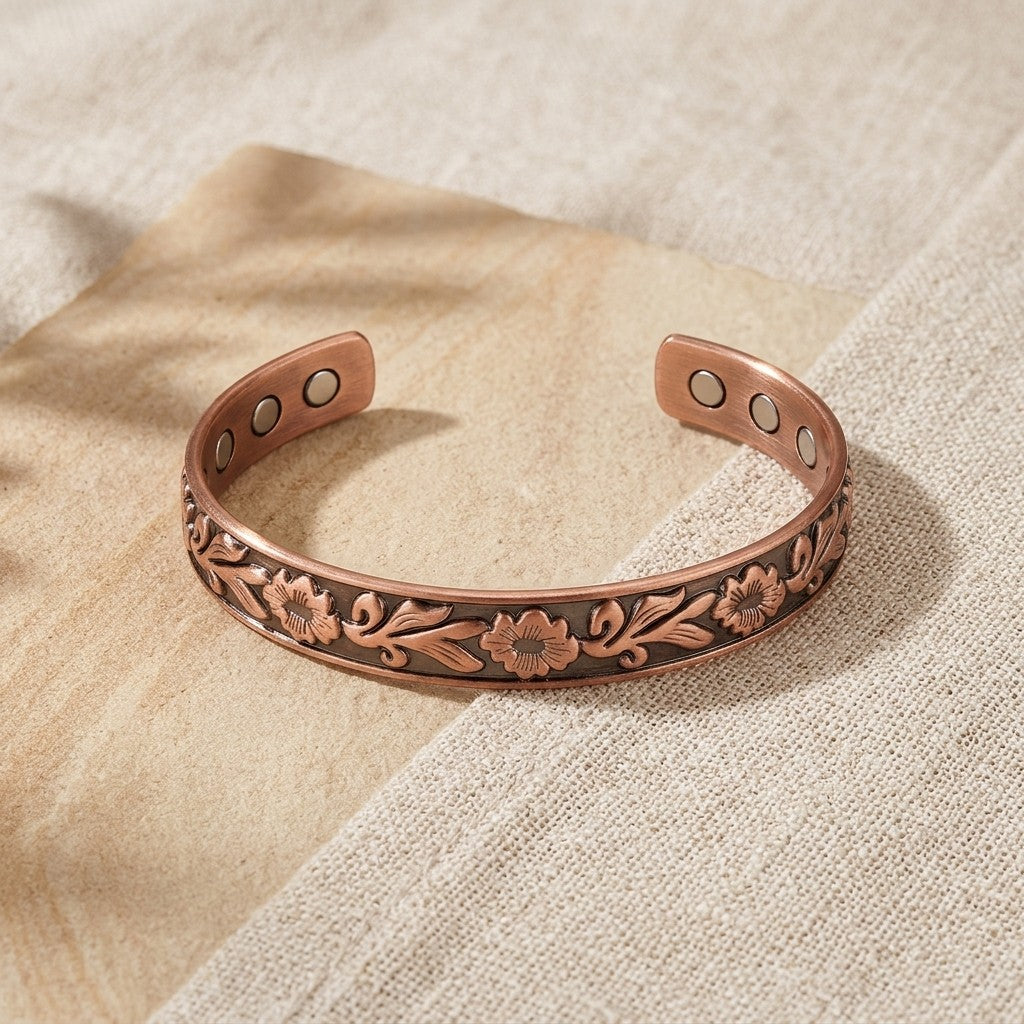 A copper cuff bracelet with embossed floral detailing and three circular magnets inserts on each side, displayed on a textured beige fabric background.