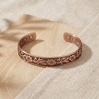 A copper cuff bracelet with embossed floral detailing and three circular magnets inserts on each side, displayed on a textured beige fabric background.