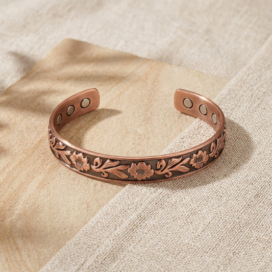 A copper cuff bracelet with embossed floral detailing and three circular magnets inserts on each side, displayed on a textured beige fabric background.