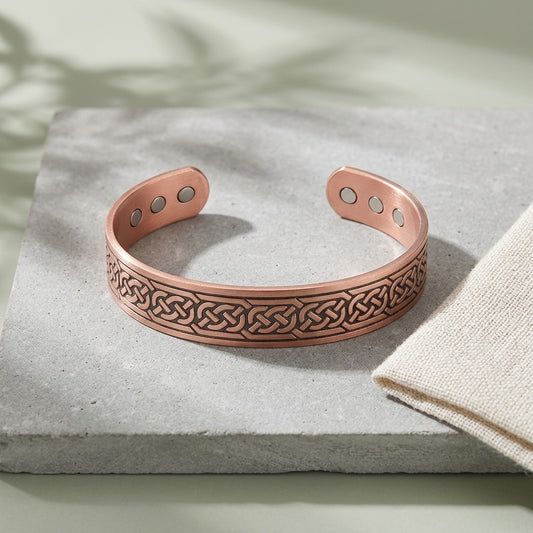 A copper magnetic cuff bracelet with an embossed Celtic knot pattern, resting on a textured grey stone slab next to a beige fabric napkin.