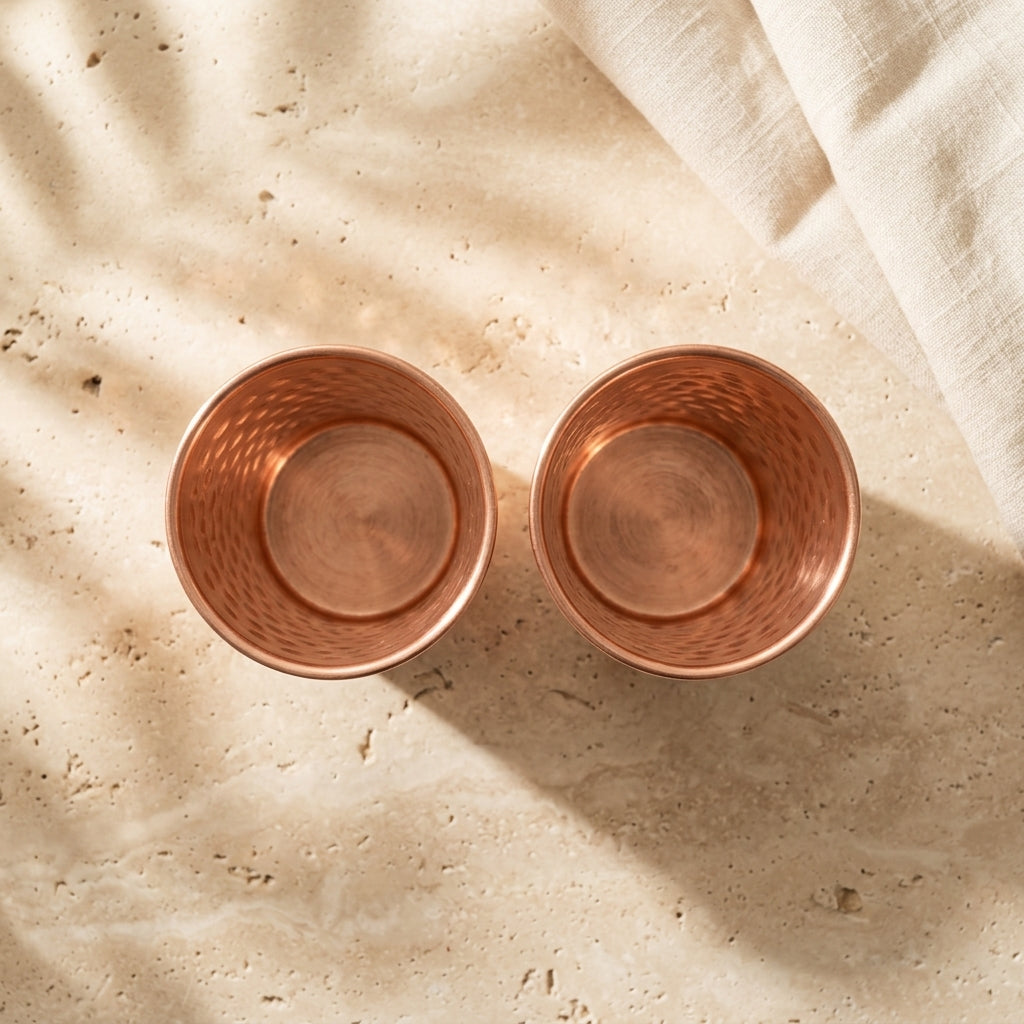 Two copper cups on a textured beige surface with a white cloth showing inner copper look