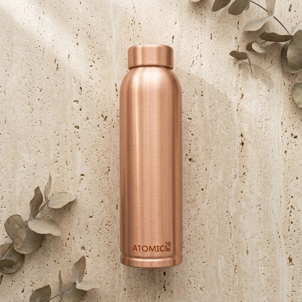 A brushed copper insulated water bottle with the brand name 'ATOMIC29' printed near the bottom, standing upright on a textured beige stone surface with scattered eucalyptus leaves.