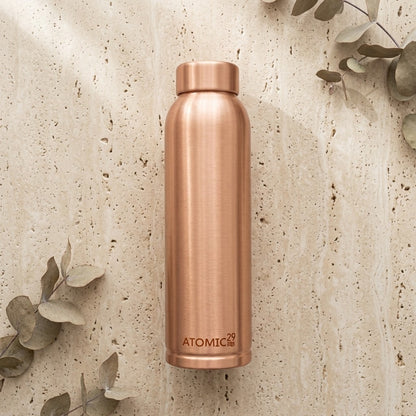 A brushed copper insulated water bottle with the brand name 'ATOMIC29' printed near the bottom, standing upright on a textured beige stone surface with scattered eucalyptus leaves.