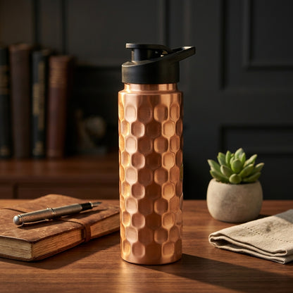 Copper water bottle with black sports cap on a wooden table with books and a plant in the background