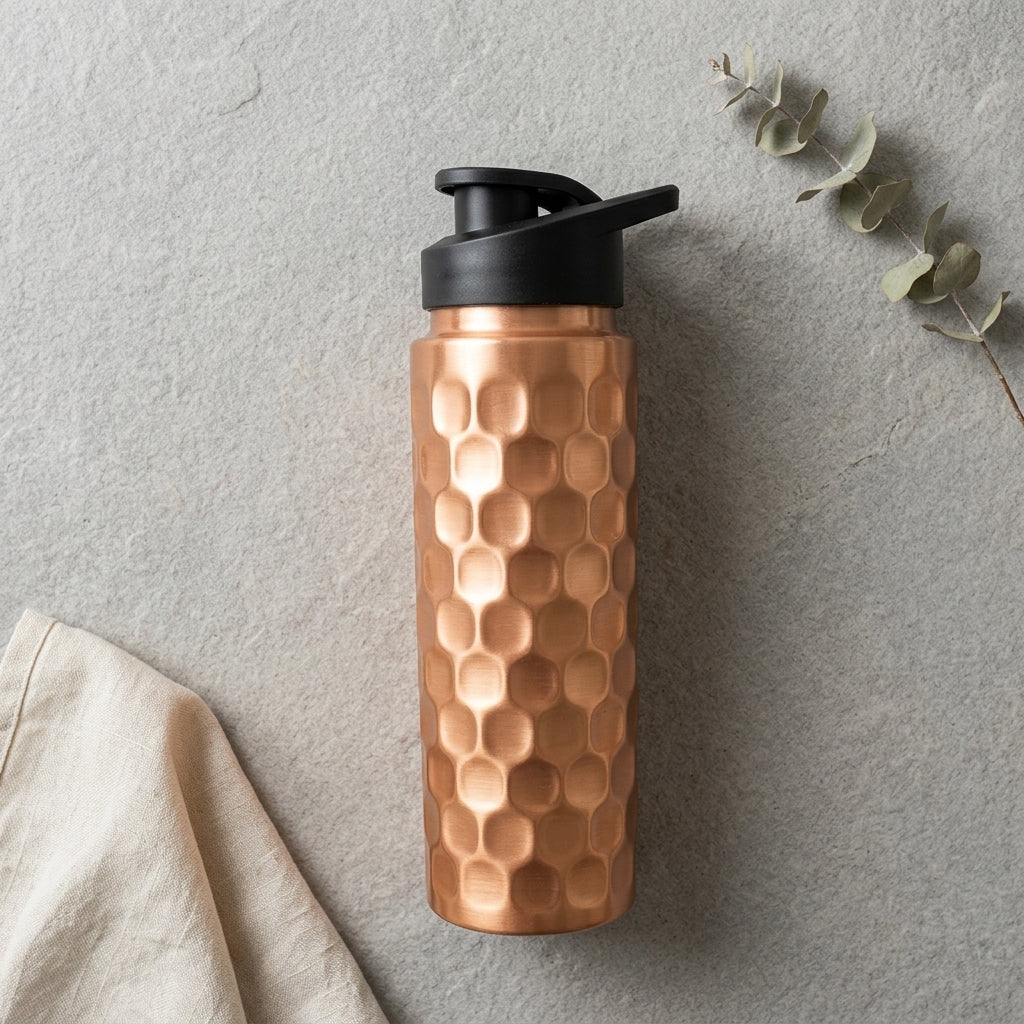 Copper water bottle with black sports cap on a grey surface with eucalyptus leaves and a beige cloth.