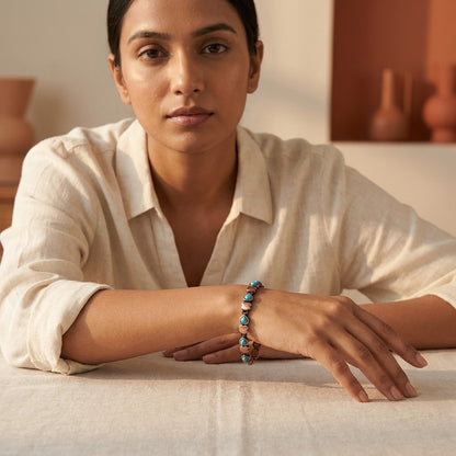 A woman wearing a cream linen shirt rests her arms on a table, showcasing aqua copper magnetic bracelet with turquoise stones inlay.