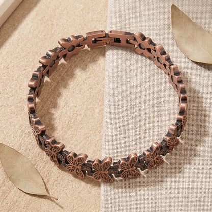 A copper butterfly link magnetic bracelet with a clasp, displayed on a split beige fabric background with dried leaves.