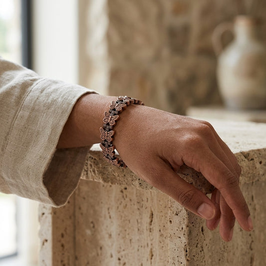 A  person's hand and forearm wearing a delicate bracelet with small copper butterfly magnetic links, resting on a stone ledge in a softly lit interior.