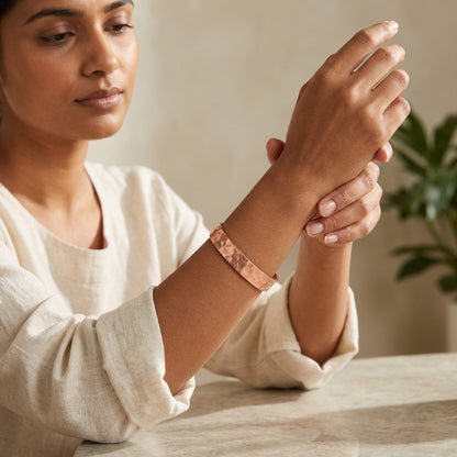 A woman wearing a hammered copper magnetic cuff bracelet, as she holds her wrist to display the bracelet. She is wearing a cream-colored linen top, and a soft-focus indoor background with a plant is visible.