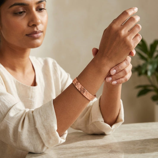 A woman wearing a hammered copper magnetic cuff bracelet, as she holds her wrist to display the bracelet. She is wearing a cream-colored linen top, and a soft-focus indoor background with a plant is visible.