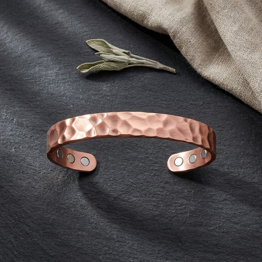 A hammered copper cuff bracelet with  magnets, displayed on a dark slate surface with a sage leaf and linen cloth in the background.
