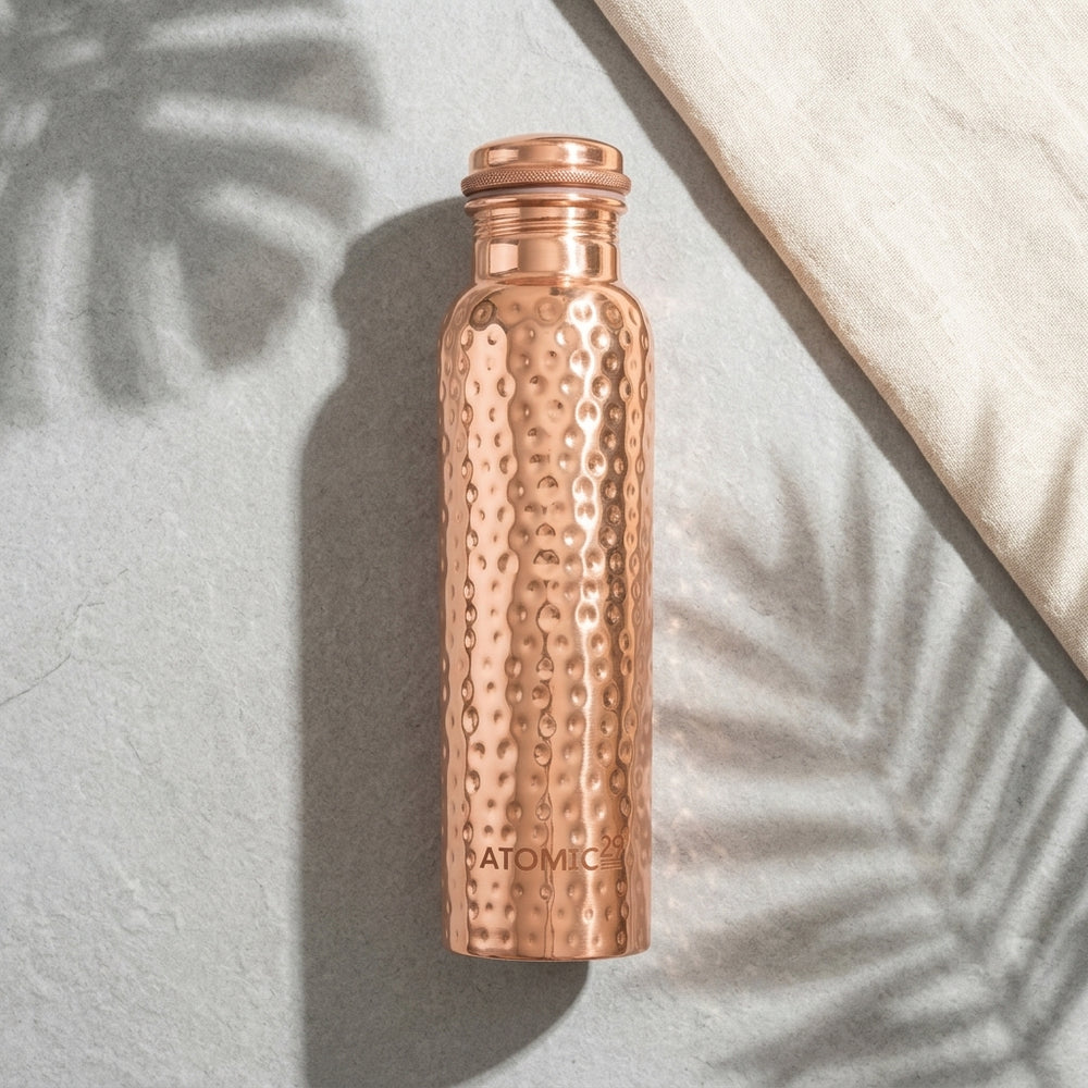 A hammered copper water bottle with a matching cap, standing upright on a textured gray surface with leaf shadows and a cream fabric corner in the background. The brand name 'ATOMIC29' is engraved near the bottom.