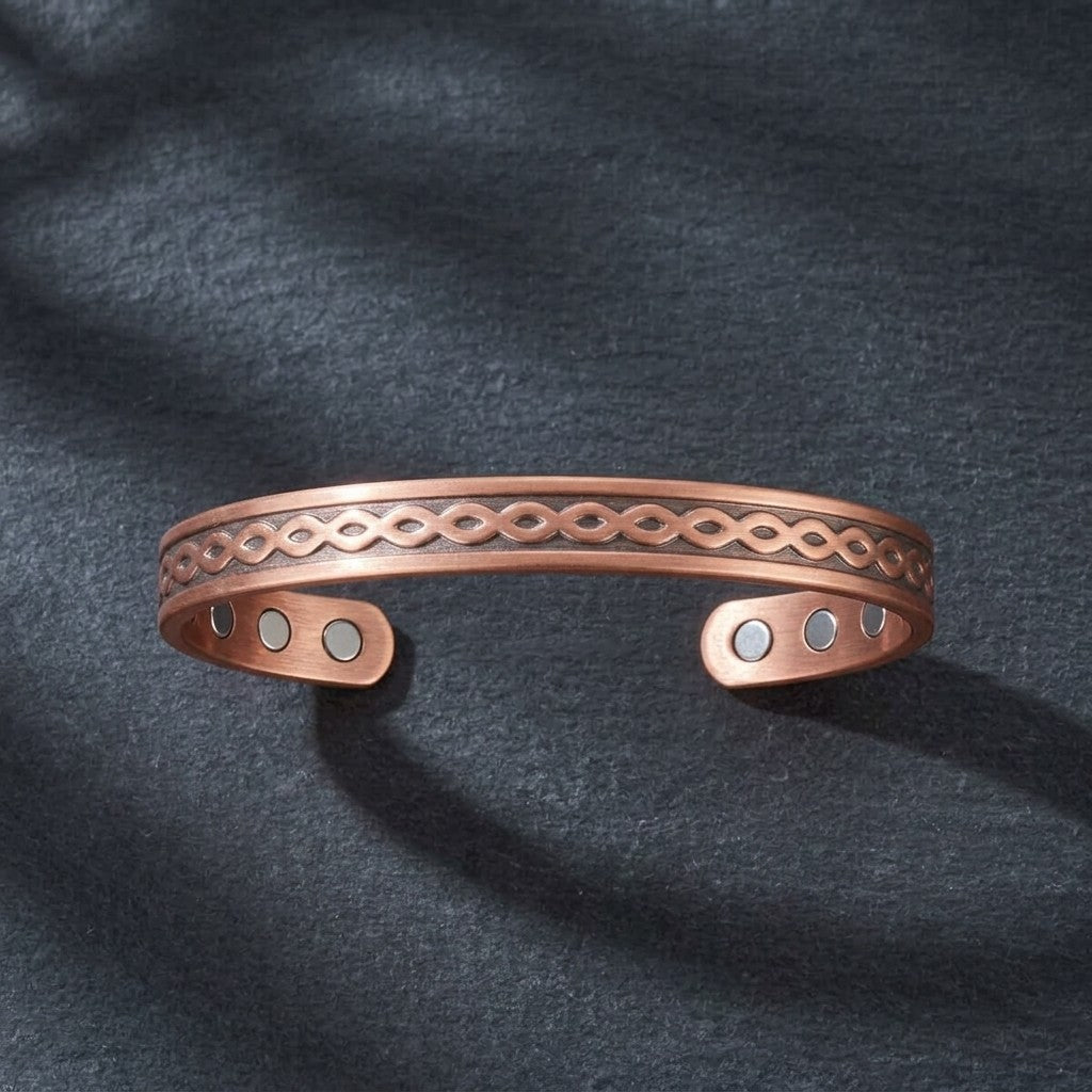 A copper open cuff bracelet with an engraved infinity pattern and three small circular magnetic accents, displayed on a dark textured fabric background.