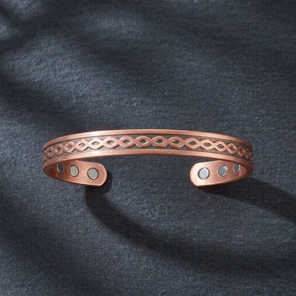 A copper open cuff bracelet with an engraved infinity pattern and three small circular magnetic accents, displayed on a dark textured fabric background.