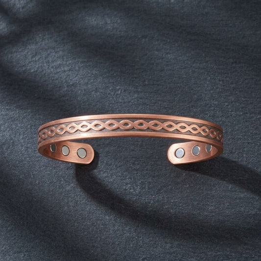 A copper open cuff bracelet with an engraved infinity pattern and three small circular magnetic accents, displayed on a dark textured fabric background.