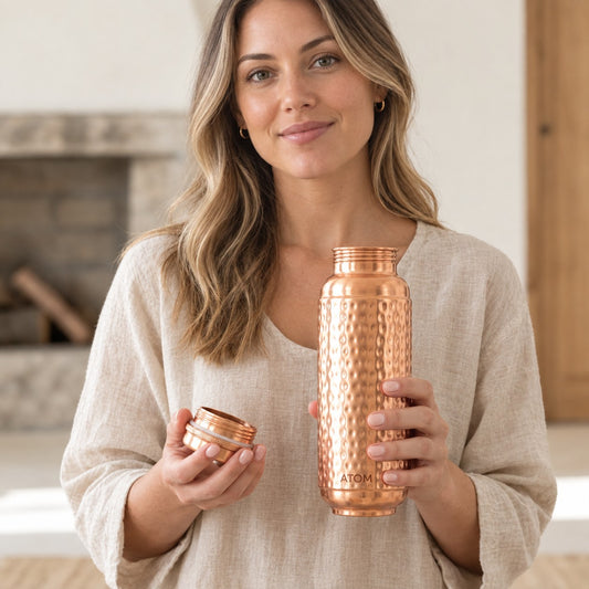 A woman in a beige linen top holds a copper water bottle and its lid in a bright, modern indoor setting. The bottle has a hammered texture and the brand name 'ATOMIC29' is visible near the base.