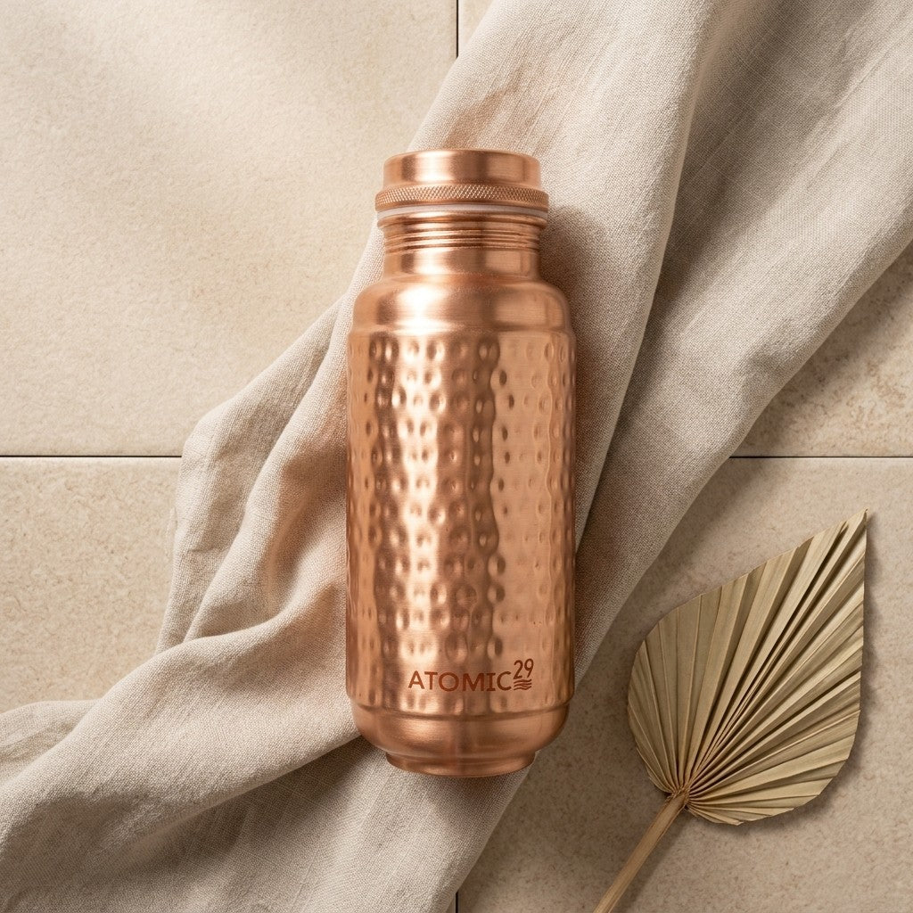 A hammered jumbo copper water bottle with the brand name 'ATOMIC29' printed near the bottom, styled on a beige fabric drape over a tiled surface with a dried palm leaf prop.
