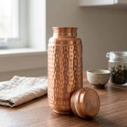 A hammered jumbo copper water bottle with its lid off, standing on a wooden table in a softly lit kitchen setting. The brand name 'ATOMIC29' is visible near the bottom of the bottle.