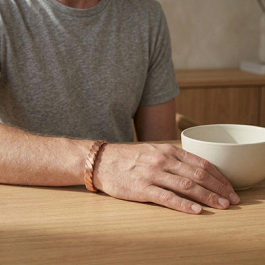 A close-up of a person's hand resting on a light wood table next to a white ceramic bowl, wearing a helix copper magnetic bracelet.