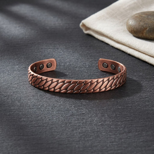 A copper magnetic cuff bracelet with a textured, embossed scale pattern, displayed on a dark Grey slate surface next to a folded beige cloth and a stone.